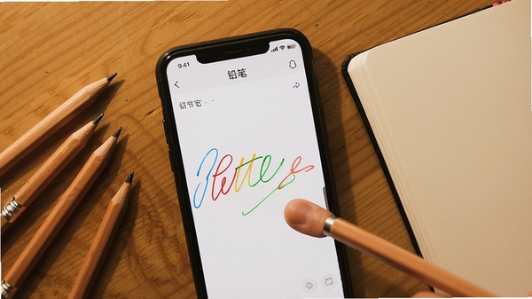 一张俯拍的智能手机屏幕特写,屏幕上正运行着名为“铅笔”的笔记软件,界面极简清爽,画面中央有用手指在触控屏上写出的流畅彩色线条笔迹,旁边散落着几支真实的木质铅笔和一本翻开的空白笔记本,暖色调桌面背景,突出高效记录与创作氛围。