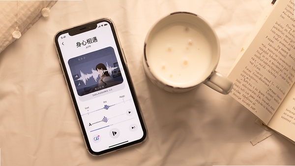 一张俯拍图,画面中一部手机正在播放“身心相遇”APP的睡眠指导音频,旁边放着一杯温热的牛奶和一本翻开的纸质书,整体色调温暖,营造出一种安详、助眠的生活化场景,暗示APP在睡眠改善方面的应用。